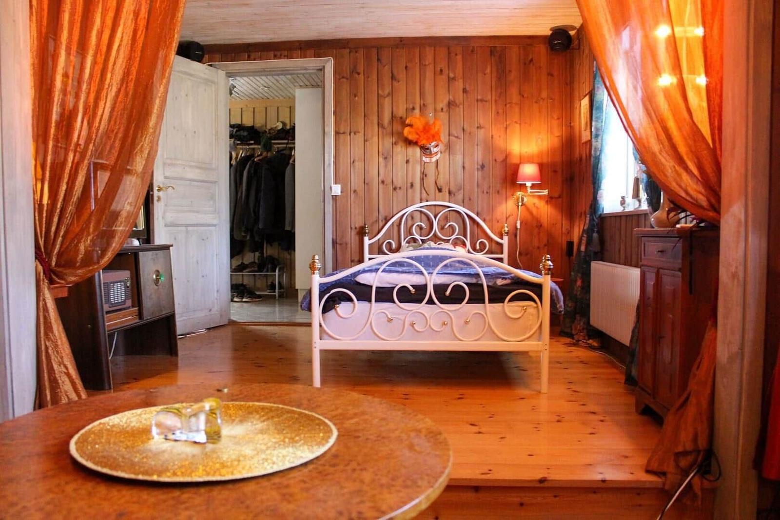 Schlafzimmer mit Holzwand, Metallbett und orangefarbenen Vorhängen.
