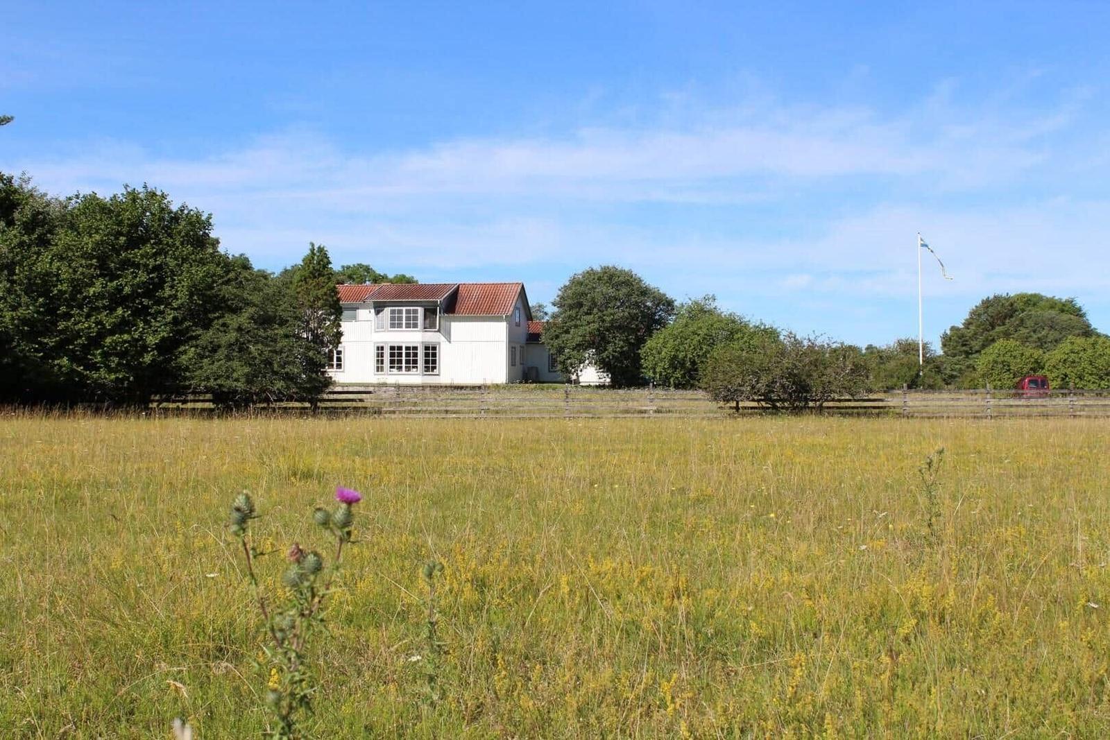 Weißes Haus mit rotem Dach, umgeben von Grasland und Bäumen unter blauem Himmel.