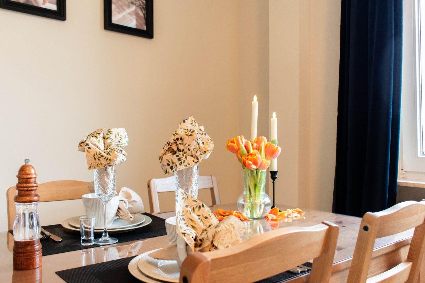 Gestellter Tisch mit Blumen, Kerzen und Geschirr. Holzstühle und dunkle Vorhänge.