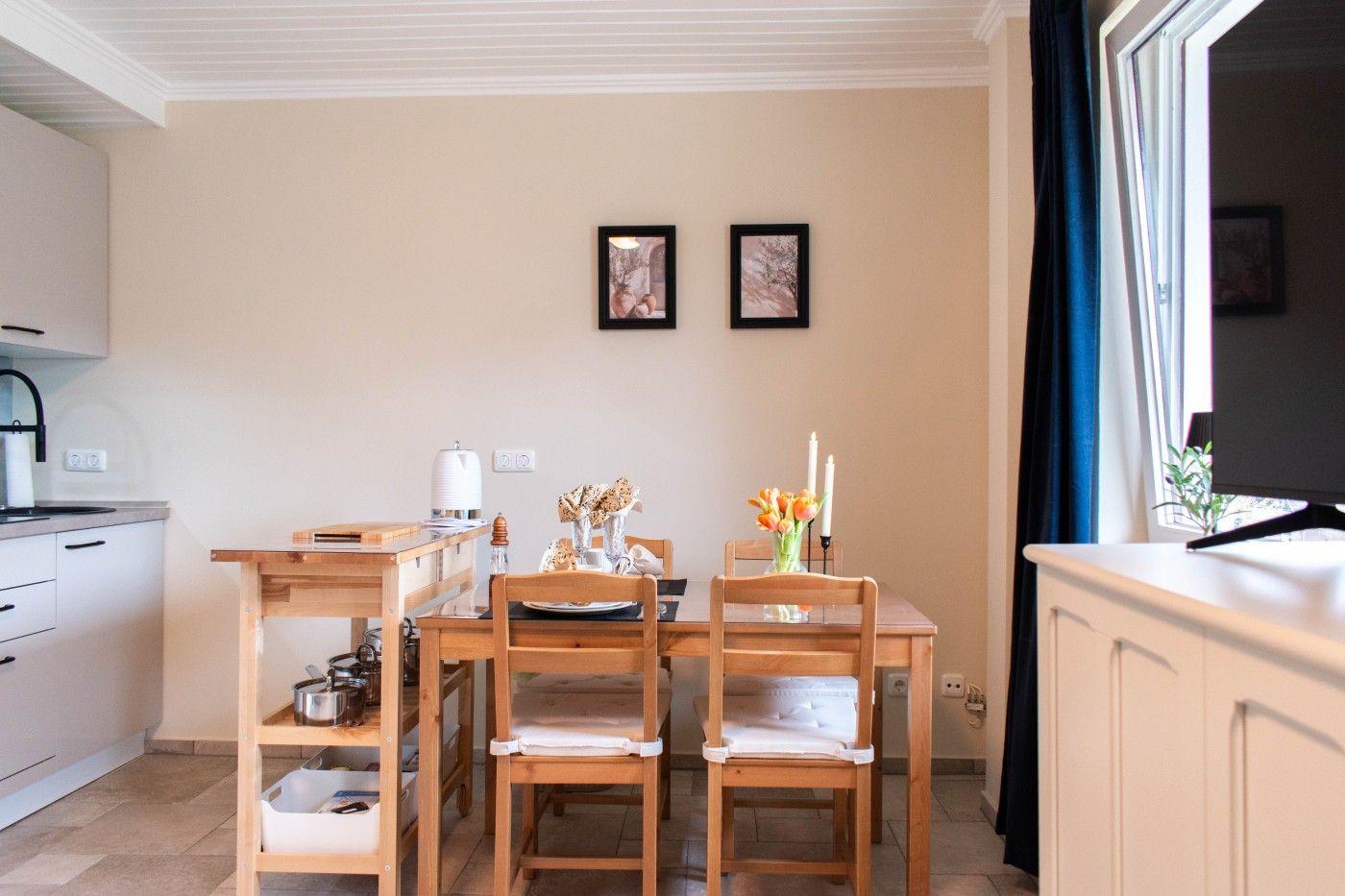 Essbereich mit Holztisch, Stühlen und Blumen. Küchenbereich und Fenster mit Vorhang sichtbar.