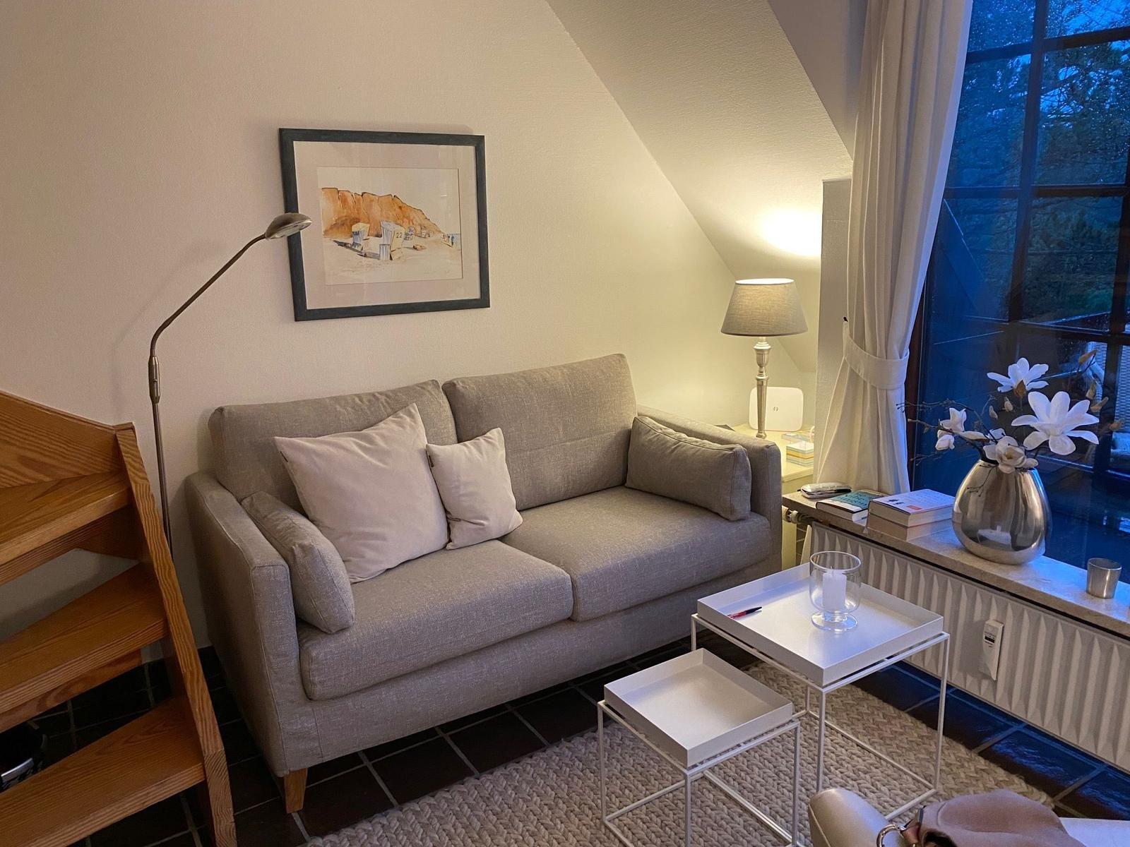 Ein Wohnbereich mit Sofa, Tischen und Fenster. Wandbild und Stehlampe sind sichtbar.