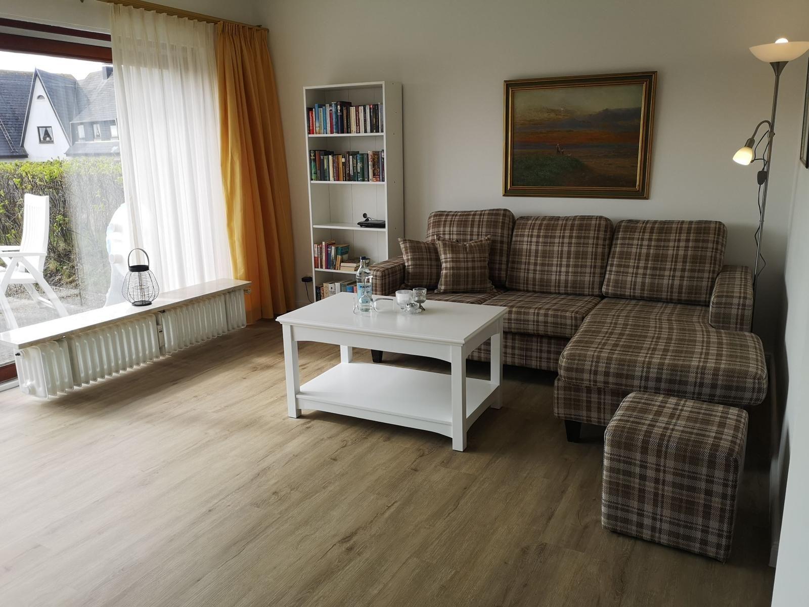 Wohnzimmer mit Sofa, Couchtisch und Bücherregal. Fenster mit Vorhängen und Blick auf Balkon.