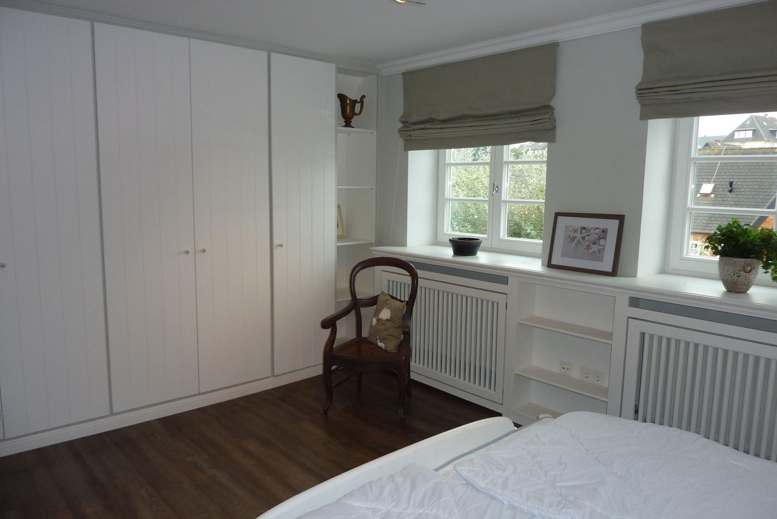 Schlafzimmer mit weißen Schränken, Holzboden und Fenstern mit Rollläden.