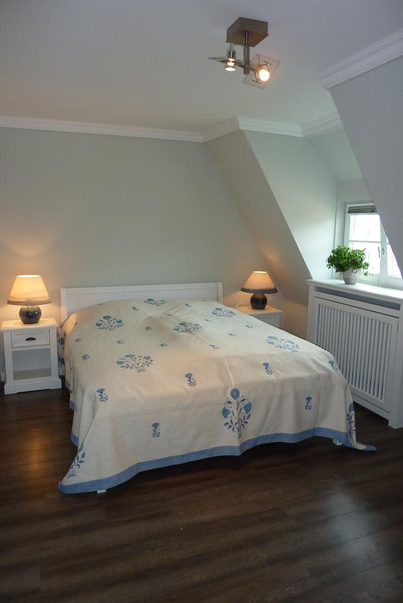 Schlafzimmer mit Dachschrägen, Bett mit blauem Bezug, zwei Nachttische und Fenster mit Pflanze.