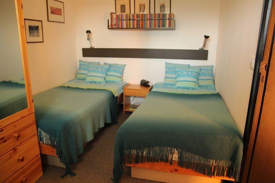Zwei Einzelbetten mit grünen Decken und Kissen. Holzkommode und Wandregal mit Büchern.