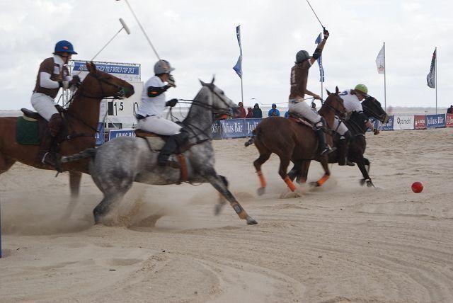Polo-Spieler reiten Pferde auf Sand und kämpfen um einen Ball.
