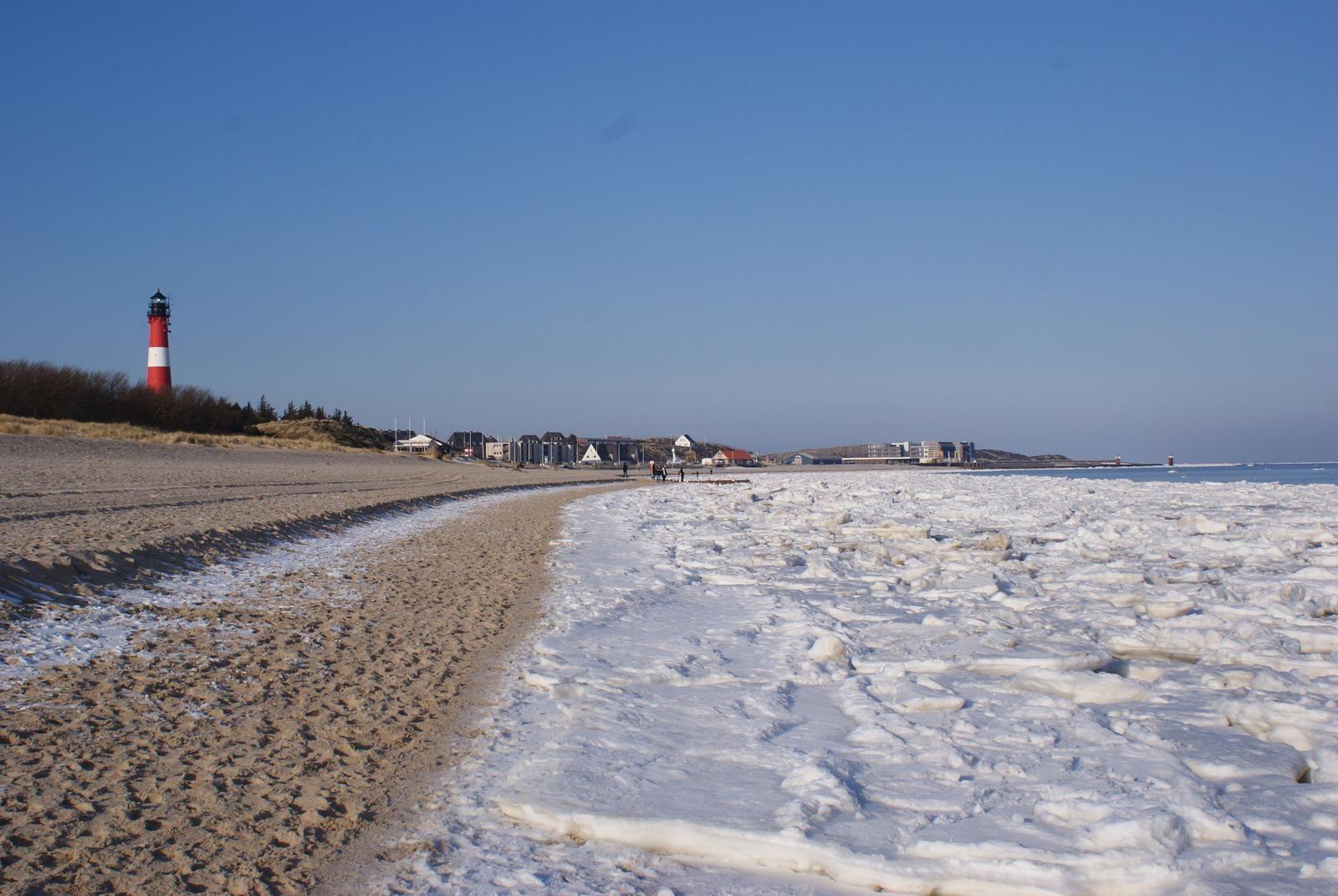 Strand mit Eis, Leuchtturm und Ferienhäusern im Hintergrund.