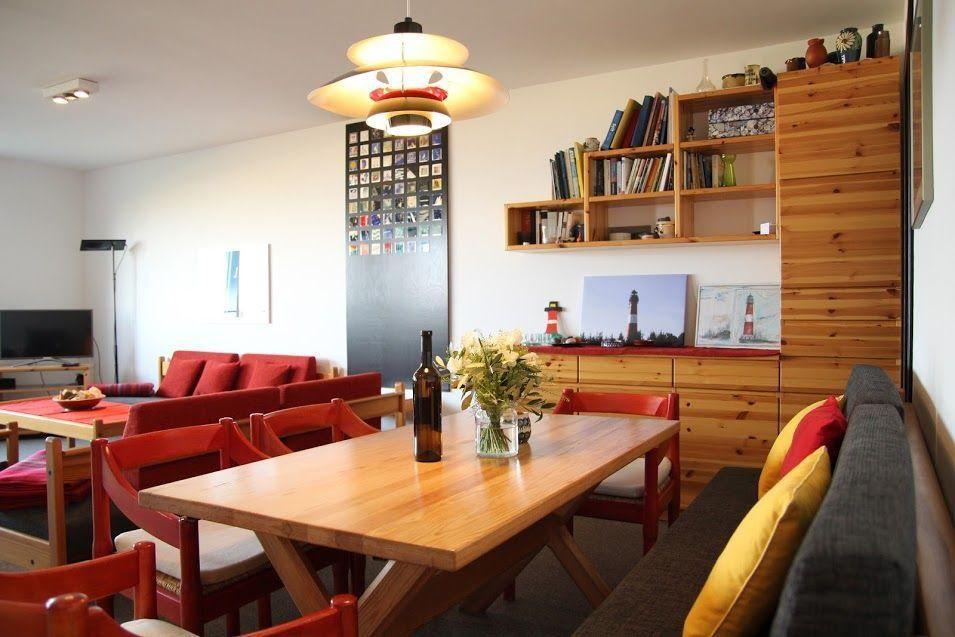 Essbereich mit Holztisch, roten Stühlen und Sofa. Wandregale und Lampen sichtbar.