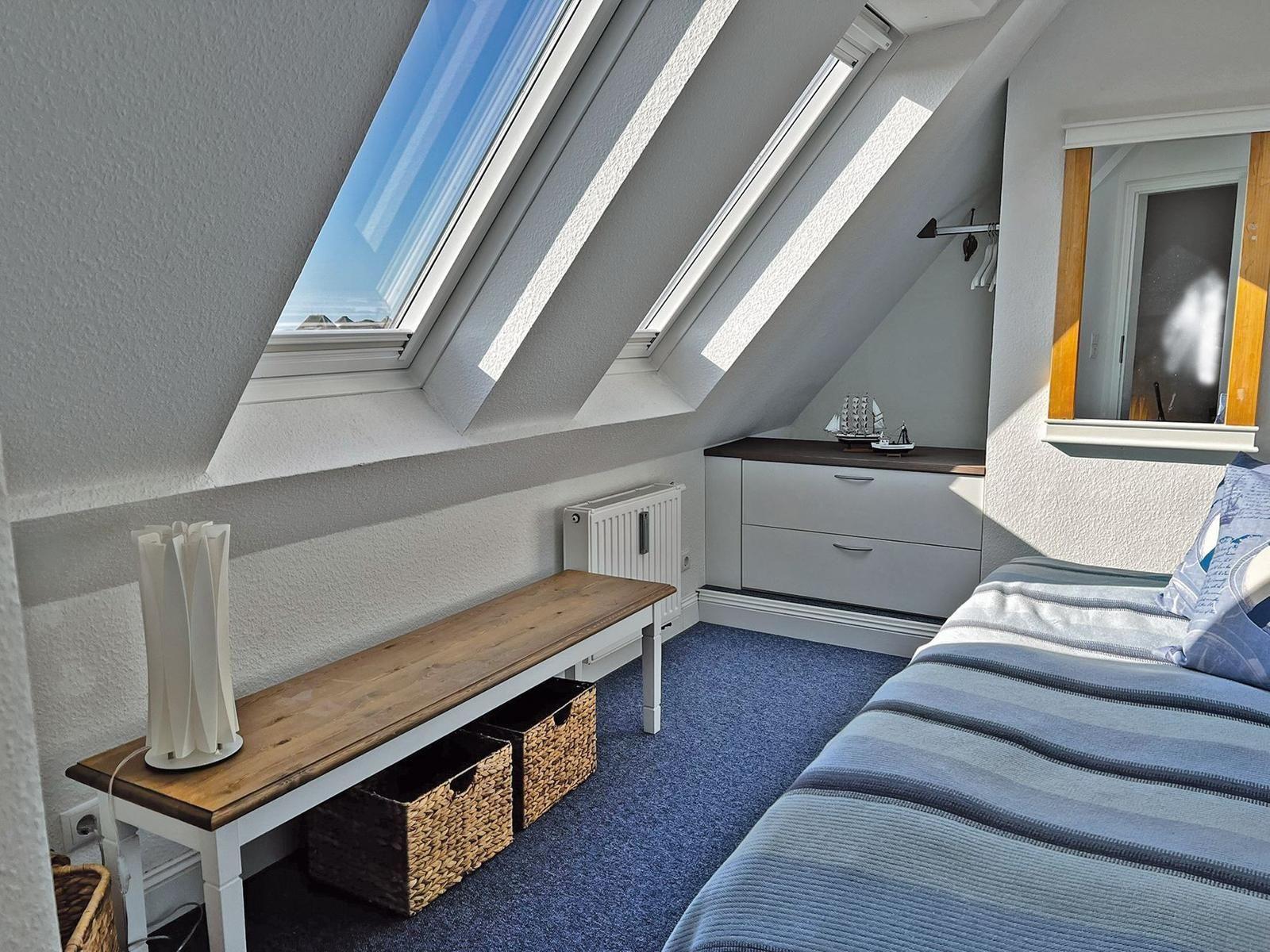 Schlafzimmer mit Dachfenstern, Bett, Schrank und Bank mit Körben.