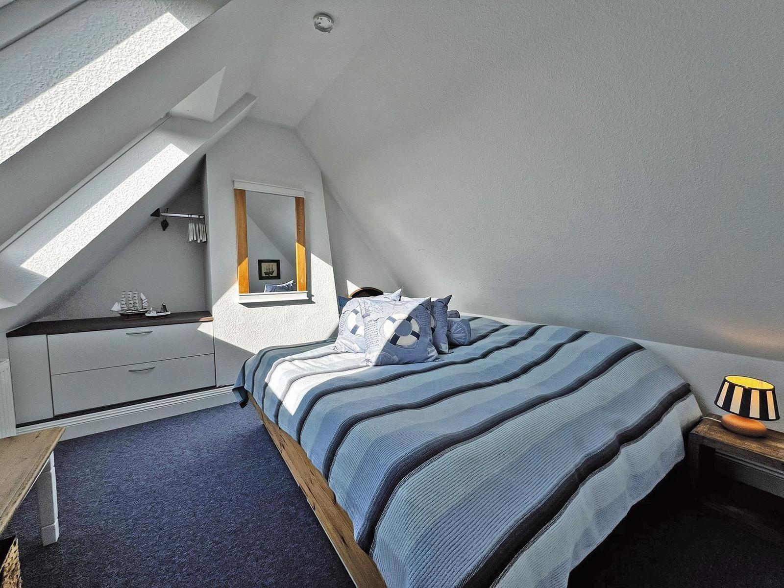 Schlafzimmer mit Dachschrägen, Bett mit blau gestreifter Bettwäsche und Kleiderschrank.
