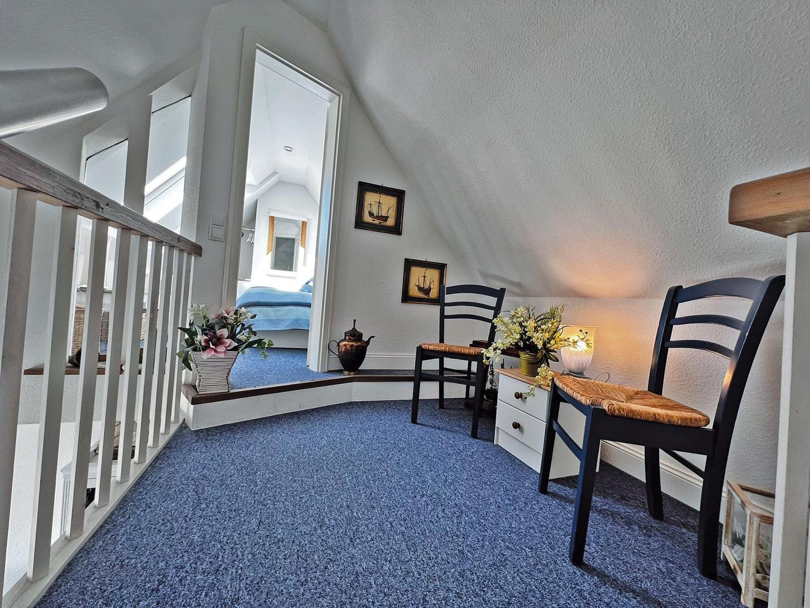 Dachgeschosszimmer mit blauem Teppich, zwei Stühlen und Blick in Schlafzimmer.