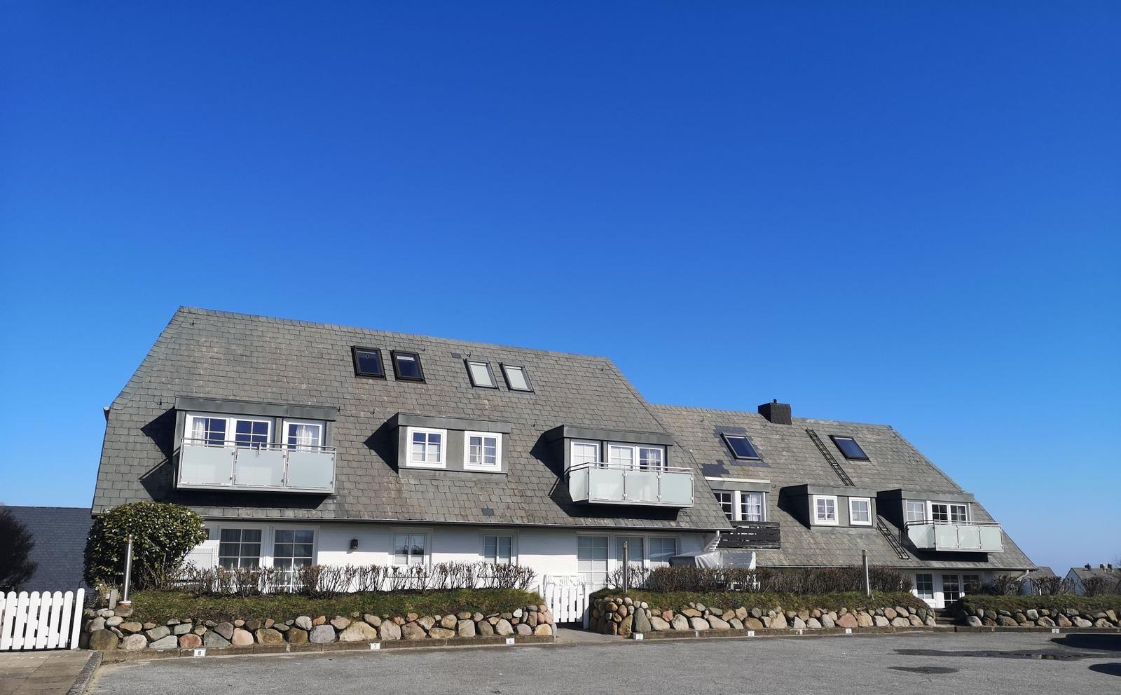 Ein zweistöckiges Haus mit grauem Dach und weißen Fenstern. Vor dem Haus befindet sich ein Steinwall.