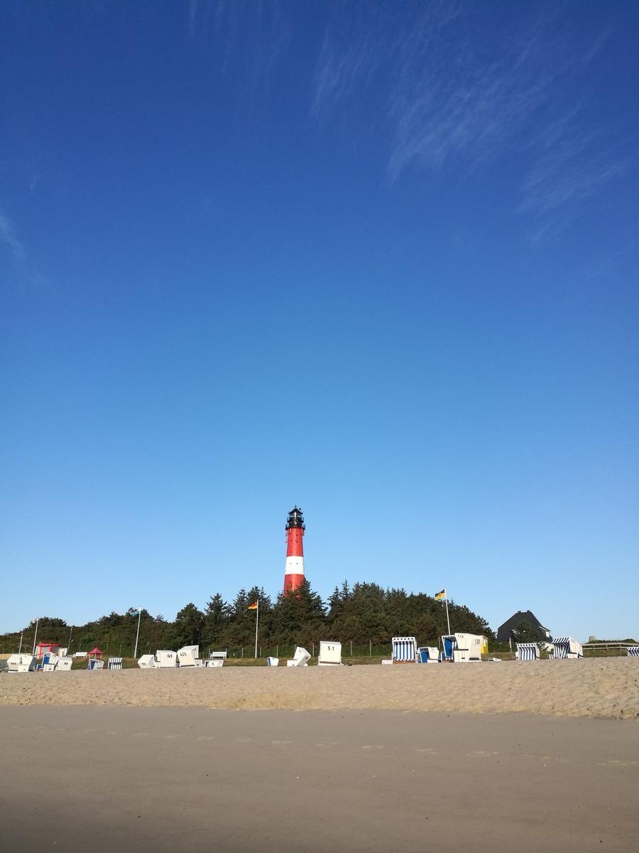 Roter Leuchtturm auf Sandstrand mit Strandkörben unter blauem Himmel.