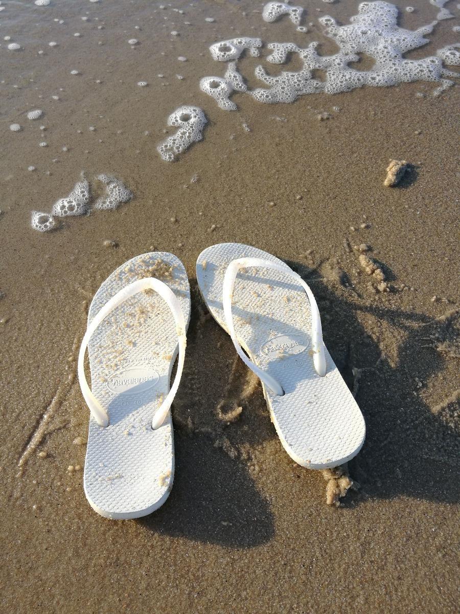 Weiße Flip-Flops liegen am sandigen Strand mit Seegras und Schaum.