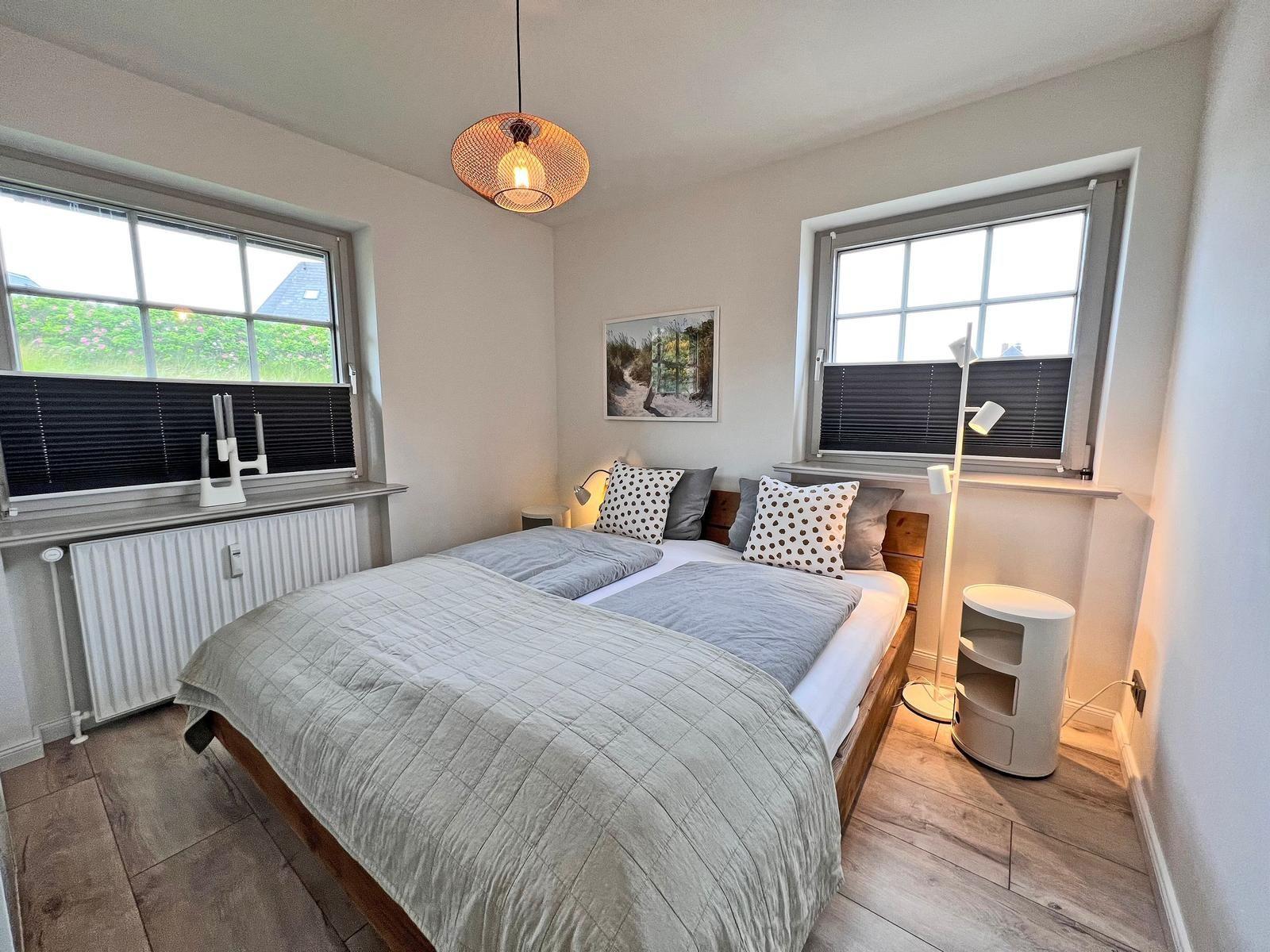 Schlafzimmer mit Doppelbett, zwei Fenstern und Deckenleuchte.