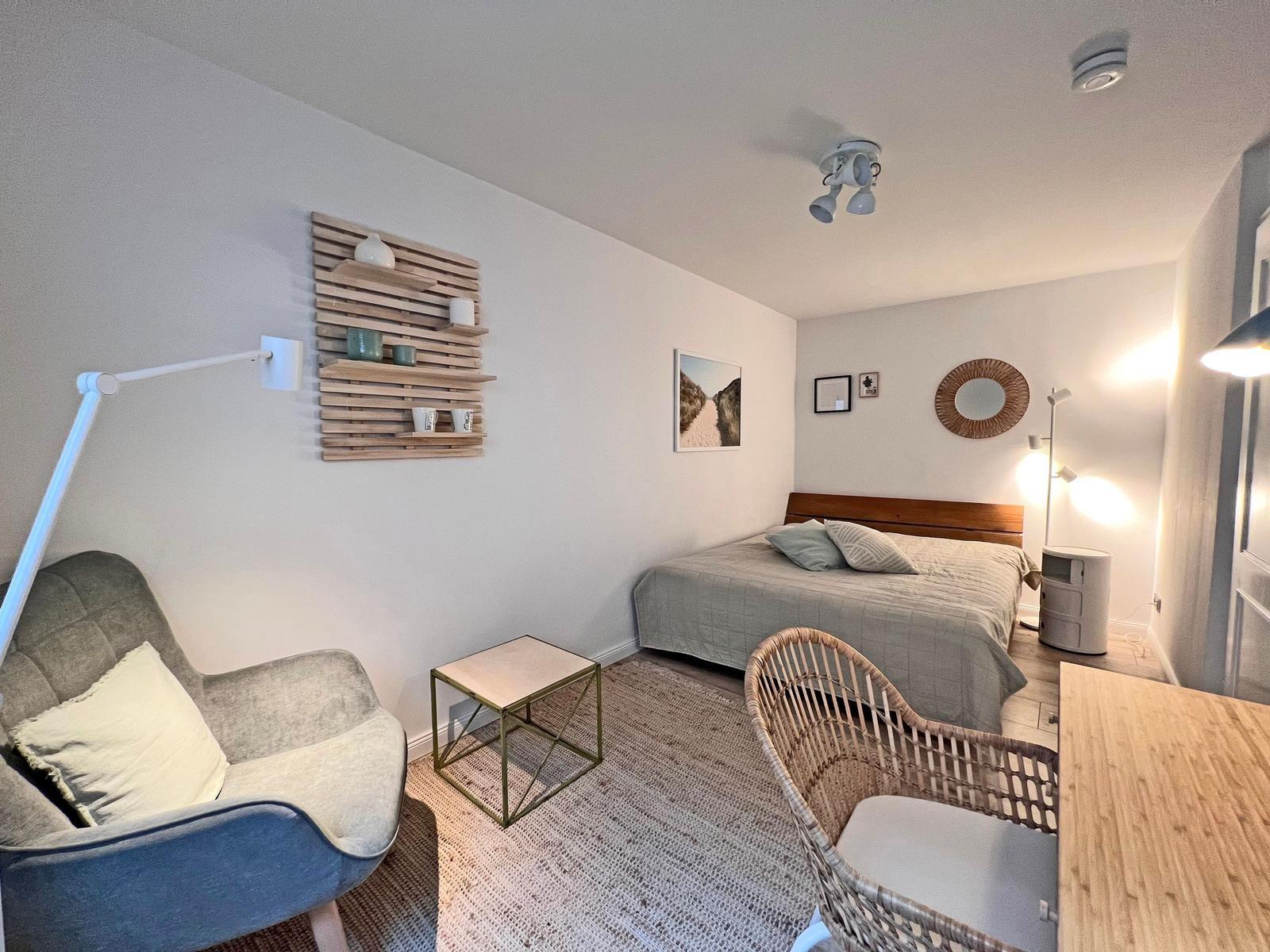 Schlafzimmer mit Bett, Sessel und Tisch. Wandregal und Wandspiegel sichtbar.