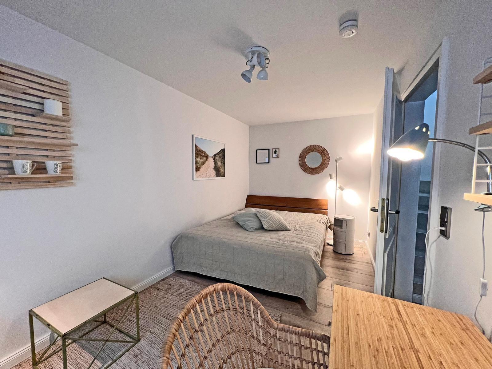 Schlafzimmer mit Bett, Wandregal und Tisch. Tür führt zu Flur.