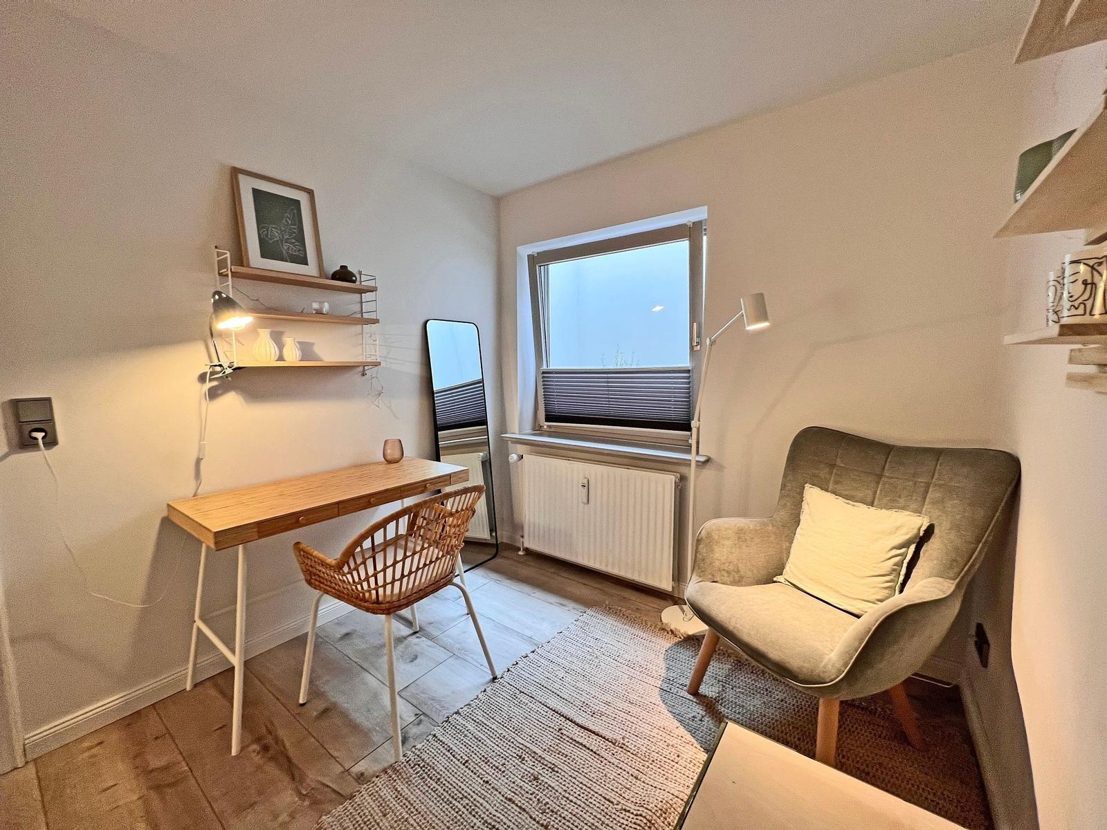 Ein Arbeitszimmer mit Schreibtisch, Stuhl und Sessel. Fenster, Wandregale und Teppich sind sichtbar.