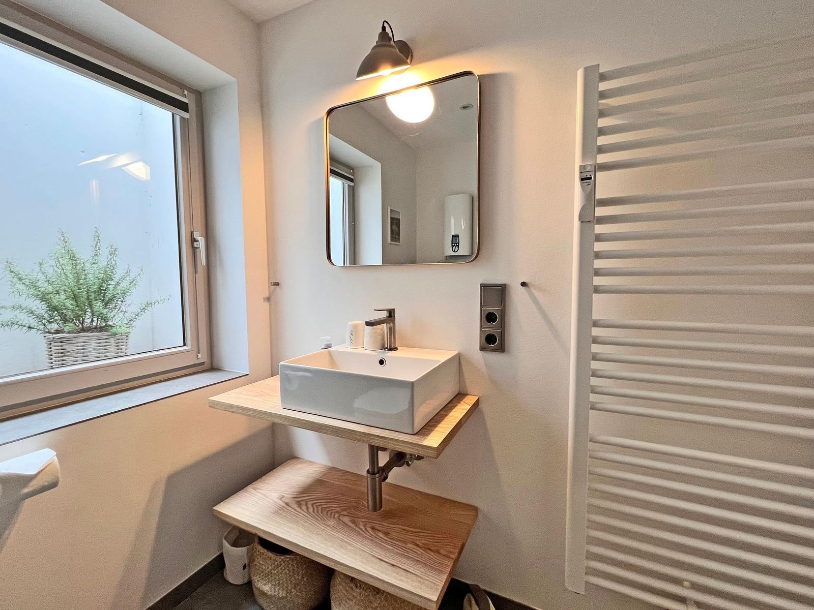 Badezimmer mit Waschbecken, Spiegel und Fenster. Links steht ein Pflanzenkorb.