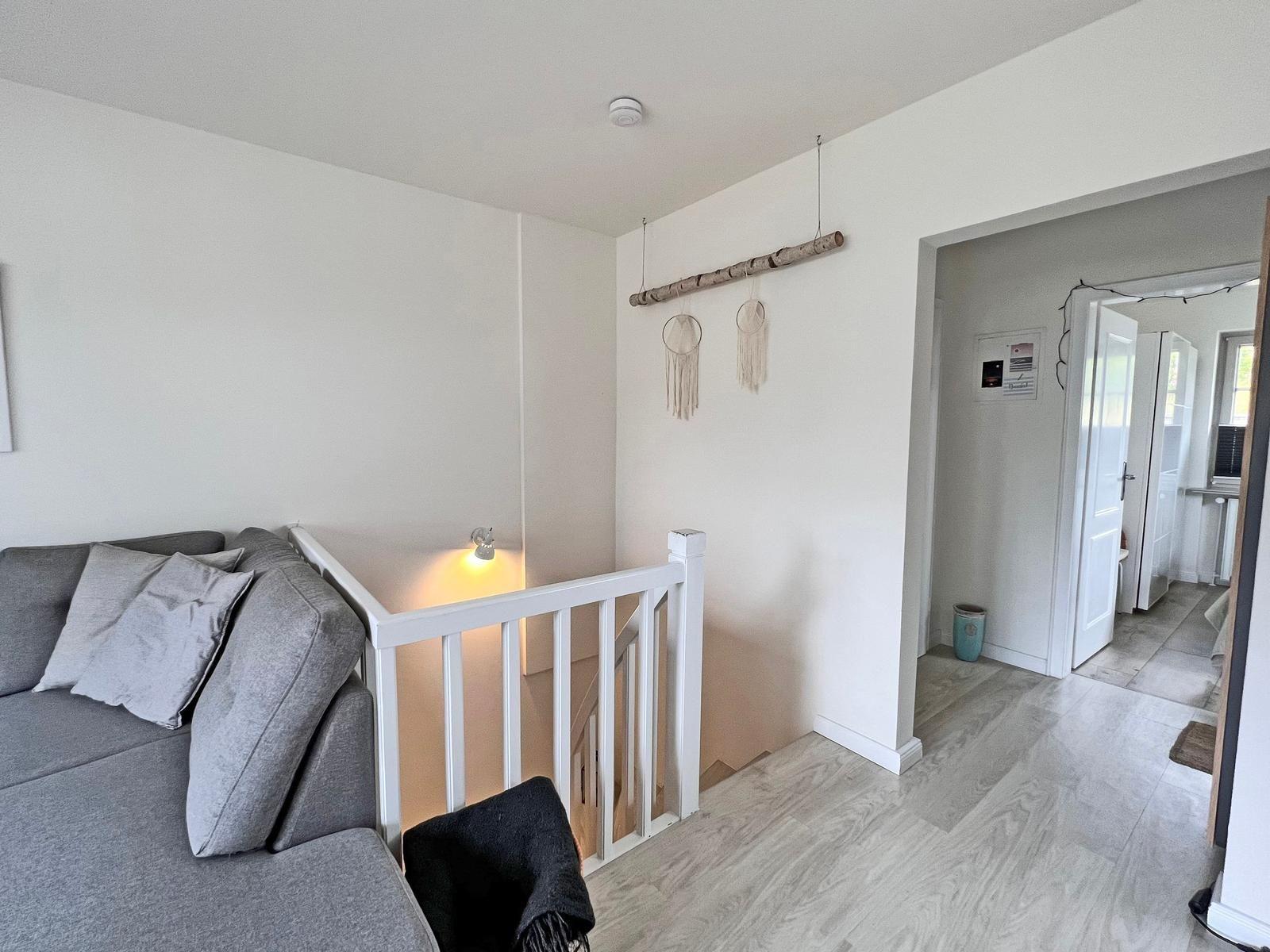 Wohnbereich mit grauem Sofa und weißen Geländern. Hintergrund zeigt einen Durchgang.