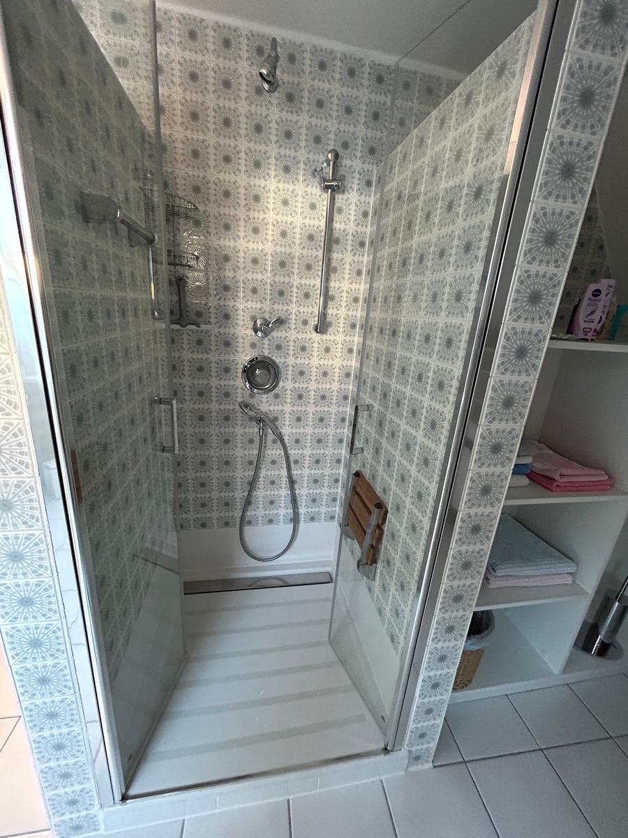 Dusche mit Glaswand und Mosaikfliesen, daneben Regal mit Handtüchern.