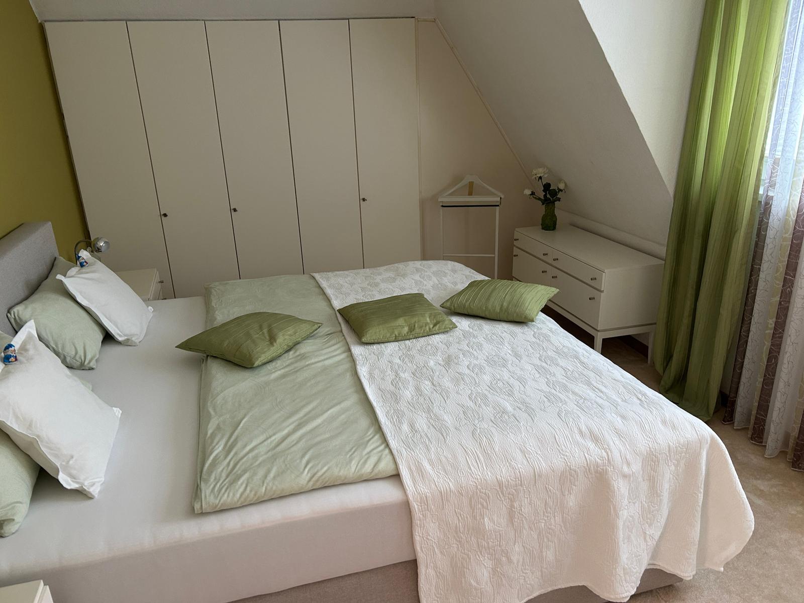 Schlafzimmer mit Doppelbett, Kleiderschrank und Fenster mit grünen Vorhängen.