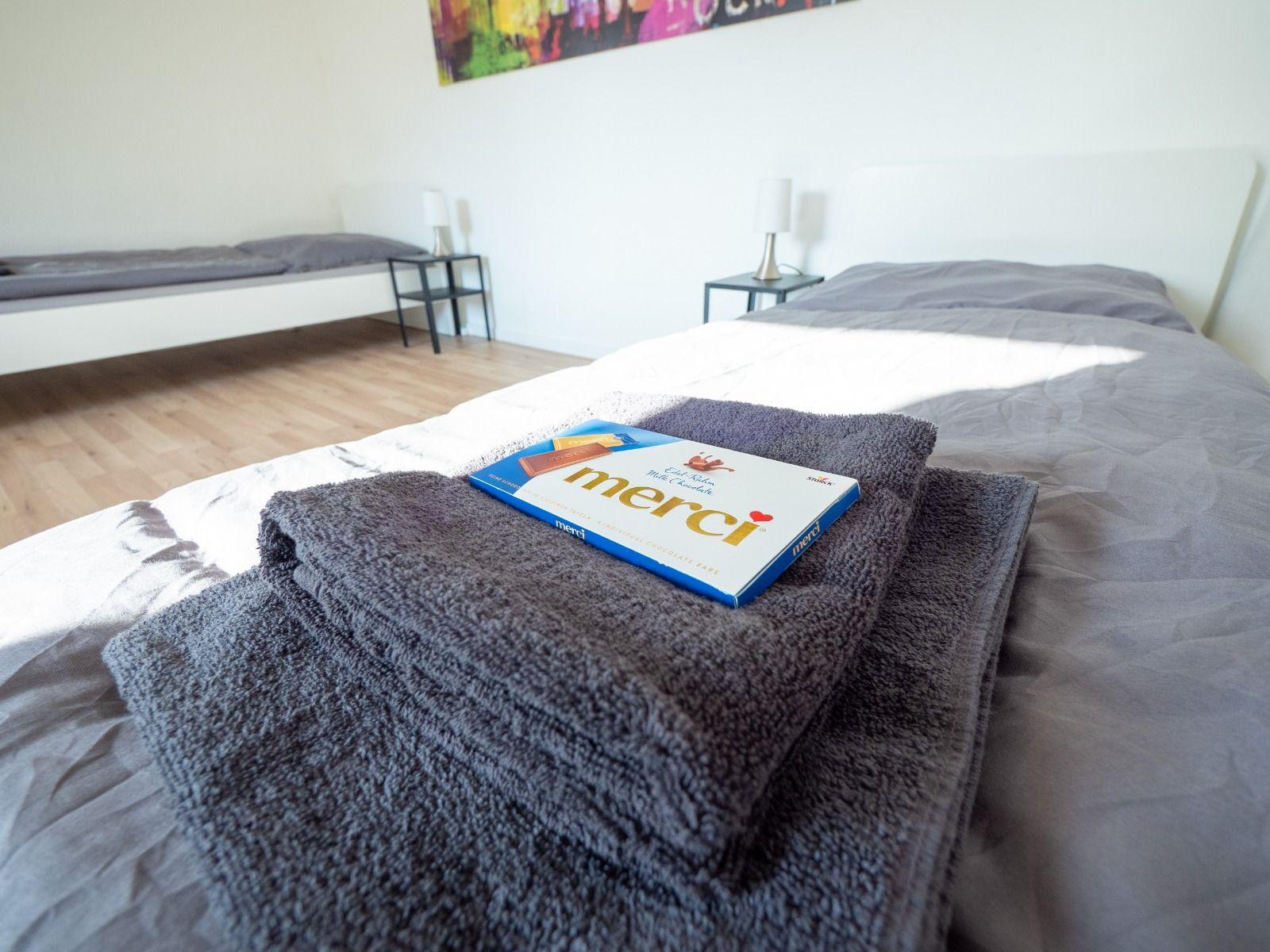 Ein Bett mit grauen Bettwäsche und einem Handtuch. Im Hintergrund ein zweites Bett und zwei Nachttische.