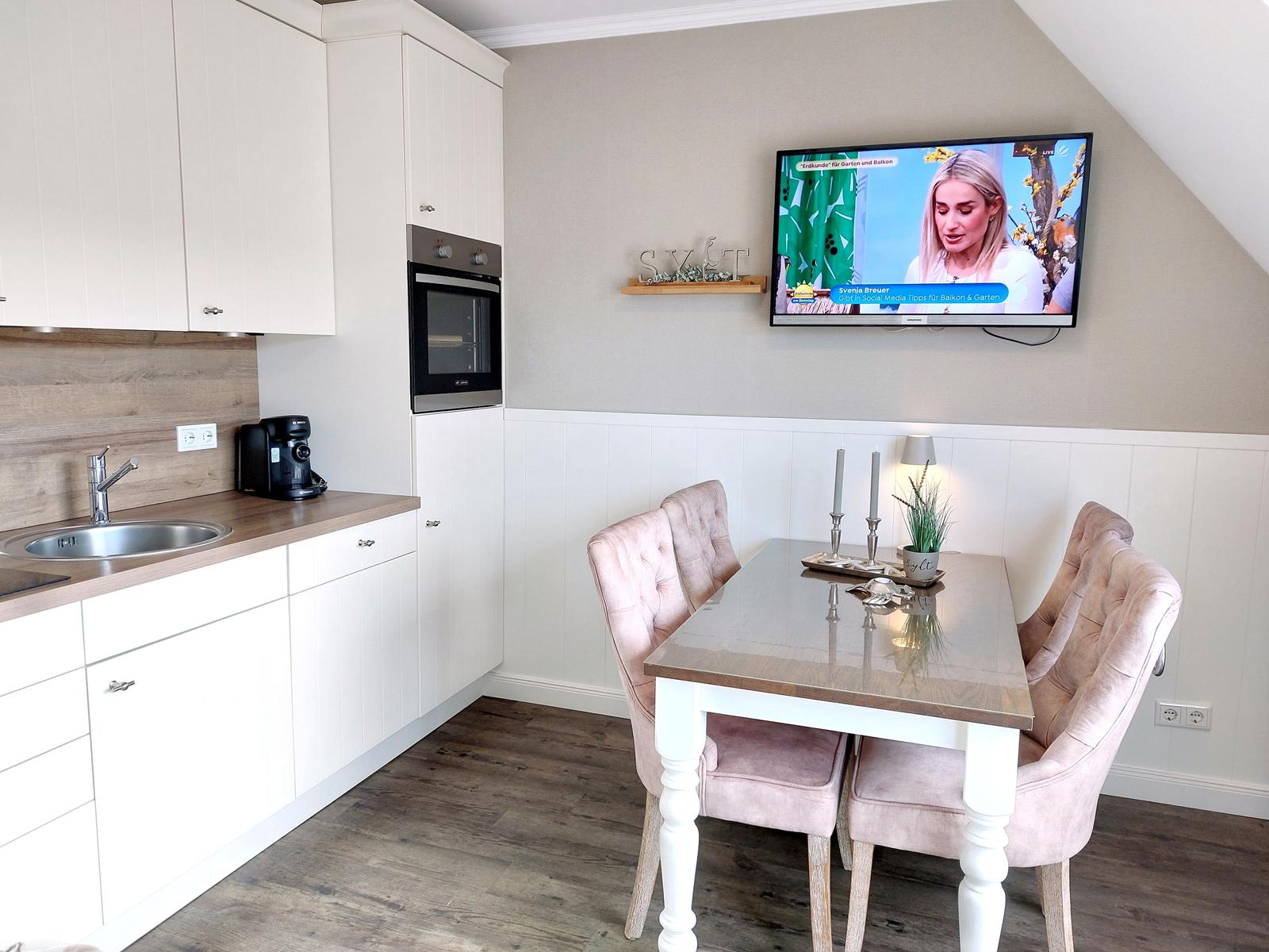 Küche mit Essbereich, TV und rosa Stühle.