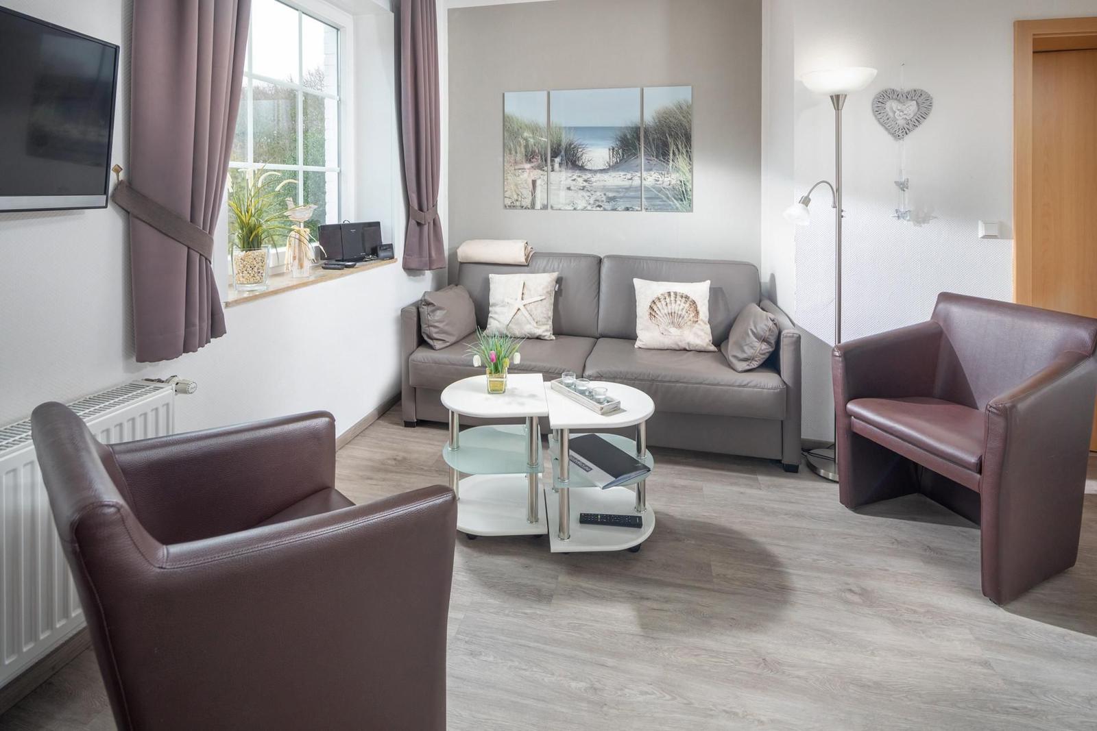 Wohnzimmer mit Sofa, zwei Sesseln und Wohnwand. Fenster mit Vorhängen und Wandbild.