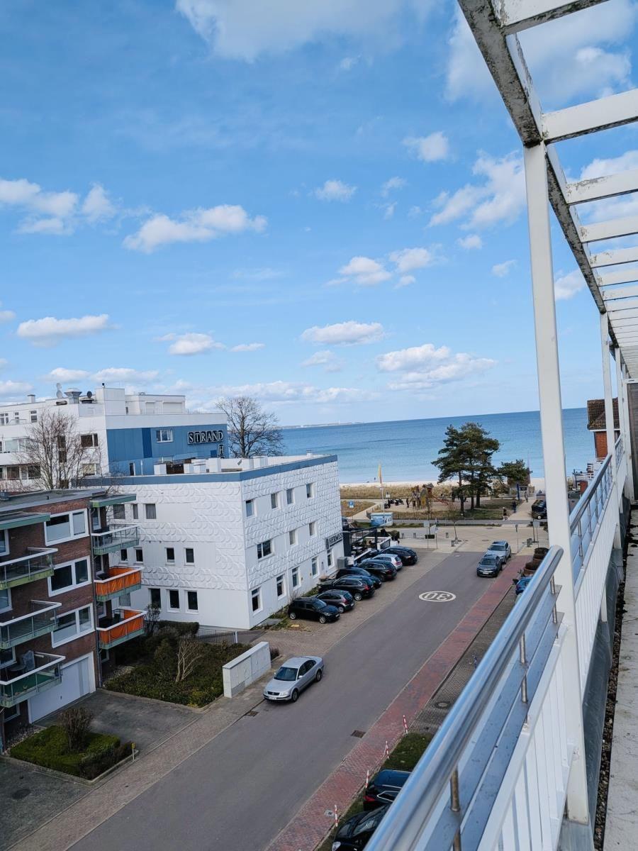Blick von Balkon auf Strand, Parkplatz und Meer unter blauem Himmel mit weißen Wolken.