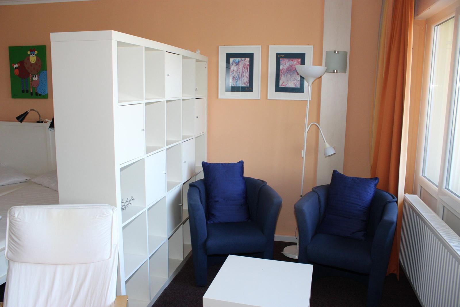 Zimmer mit zwei blauen Sesseln, weißem Tisch und Regal. Fenster mit orangefarbenen Vorhängen.