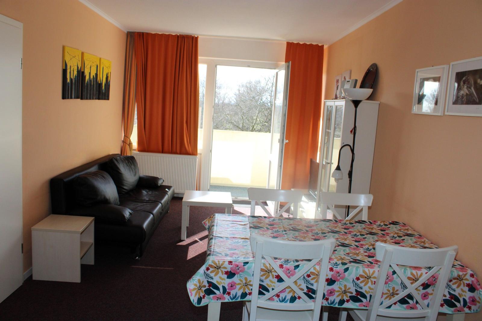 Wohn- und Essbereich mit Sofa, Tisch und Stühlen, orangefarbene Vorhänge und Wandbilder.