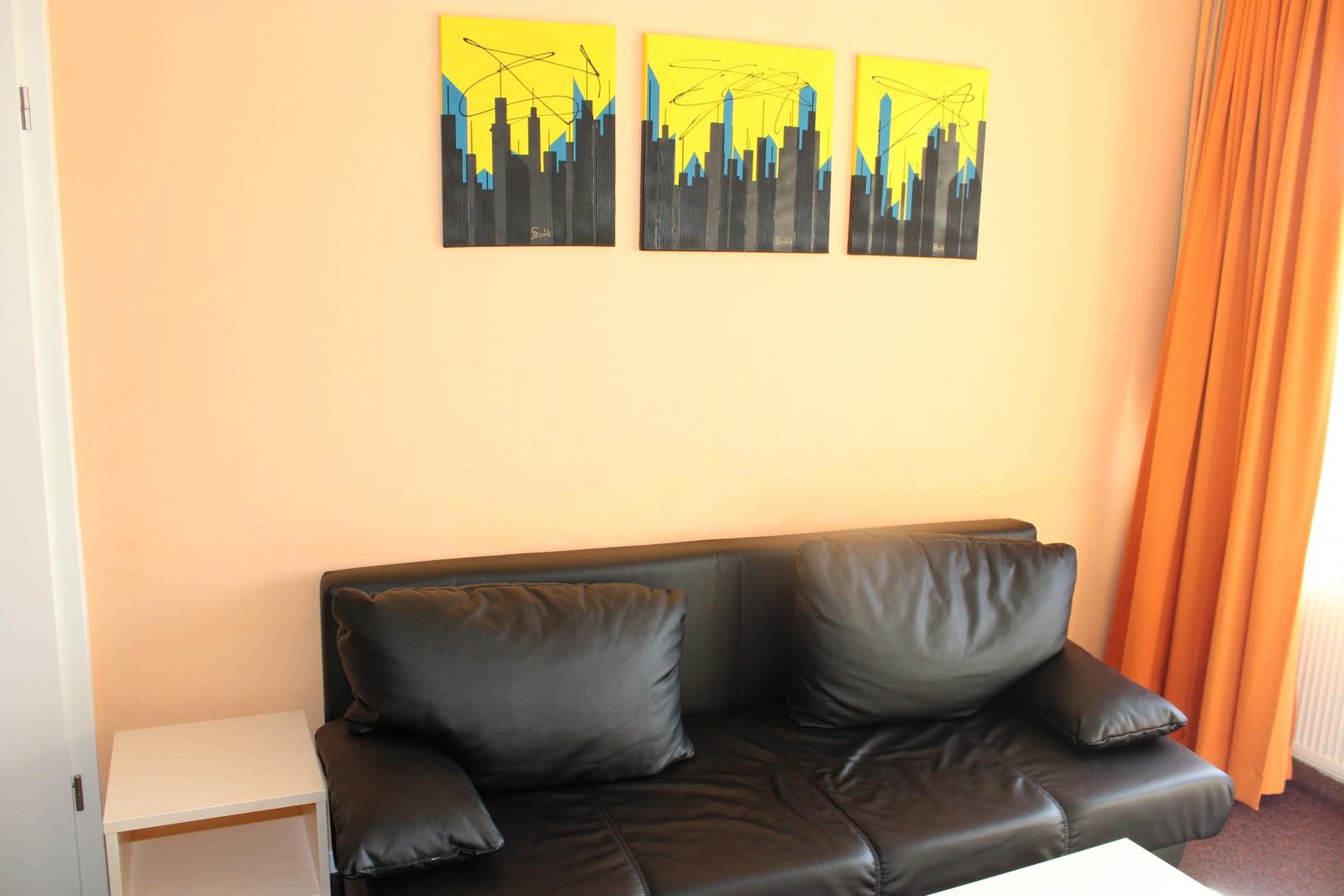 Schwarzes Sofa mit Kissen, Wand mit drei Kunstwerken, orange Vorhänge.
