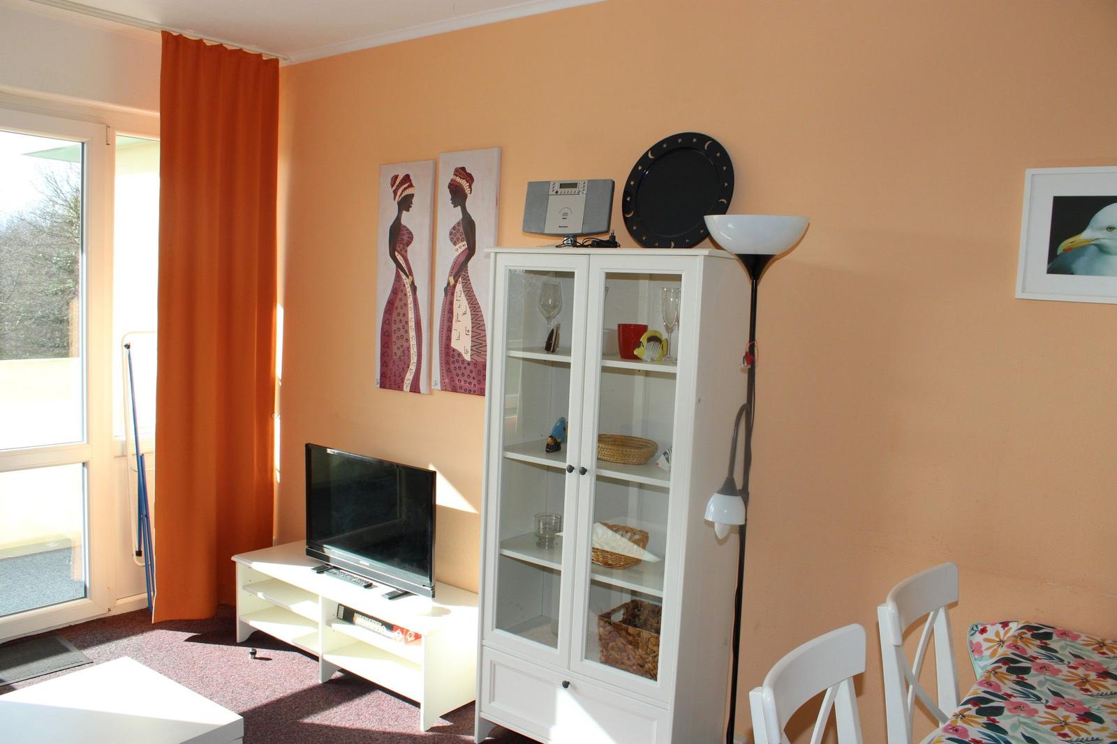 Wohnzimmer mit Fernseher, Glasvitrine und orangefarbenen Vorhängen.