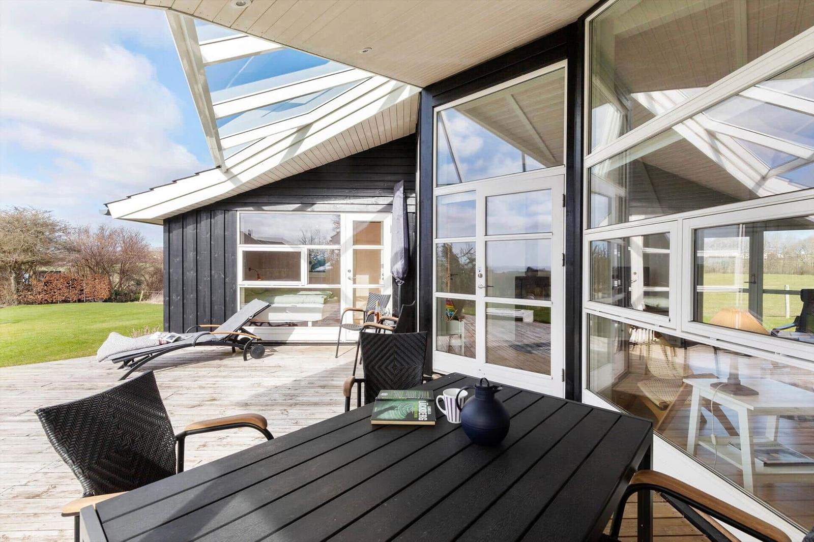 Terrasse mit Tisch, Stühlen und Liegestuhl. Glasfront und Dach überdacht.