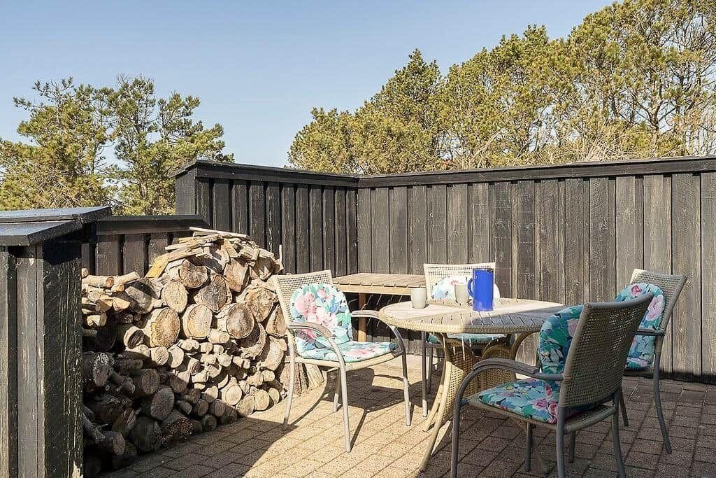 Terrasse mit Tisch, Stühlen und Holzstapel. Hintergrund: dunkler Holzzaun und Bäume.