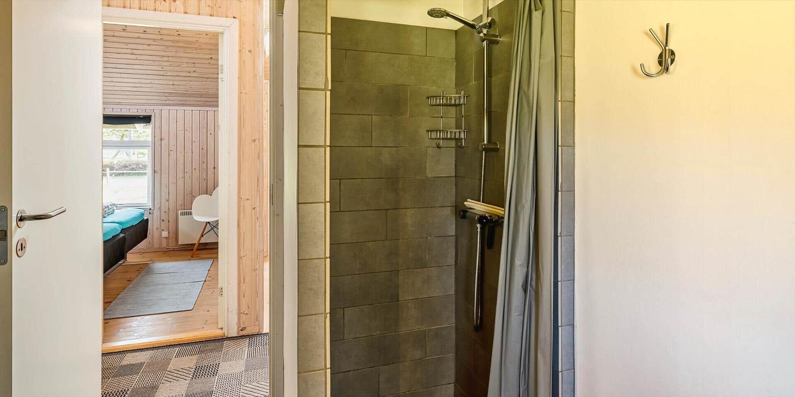 Dusche mit Duschbrause und Regal. Tür führt in Schlafzimmer mit Bett und Fenster.