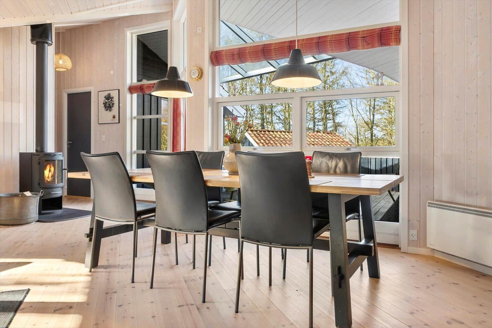 Esszimmer mit Holztisch, schwarzen Stühlen und Kamin. Große Fenster mit Blick auf die Natur.