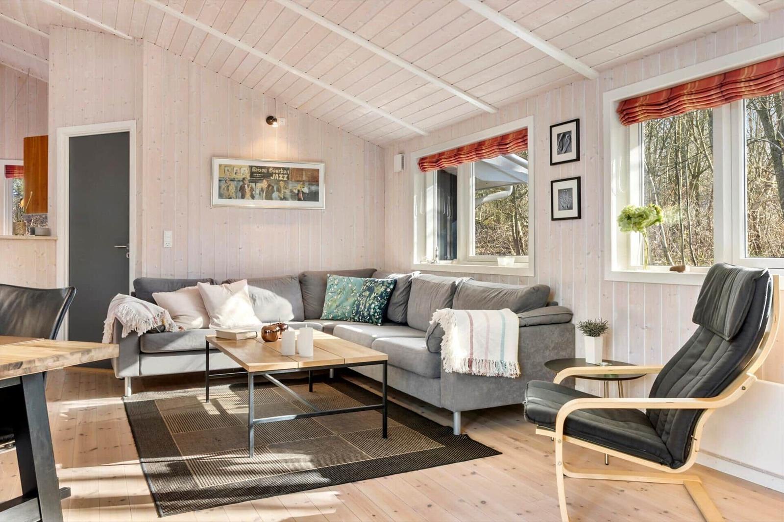 Wohnzimmer mit grauem Sofa, Holztisch und Sessel. Holzwände und Fenster mit Blick in den Wald.