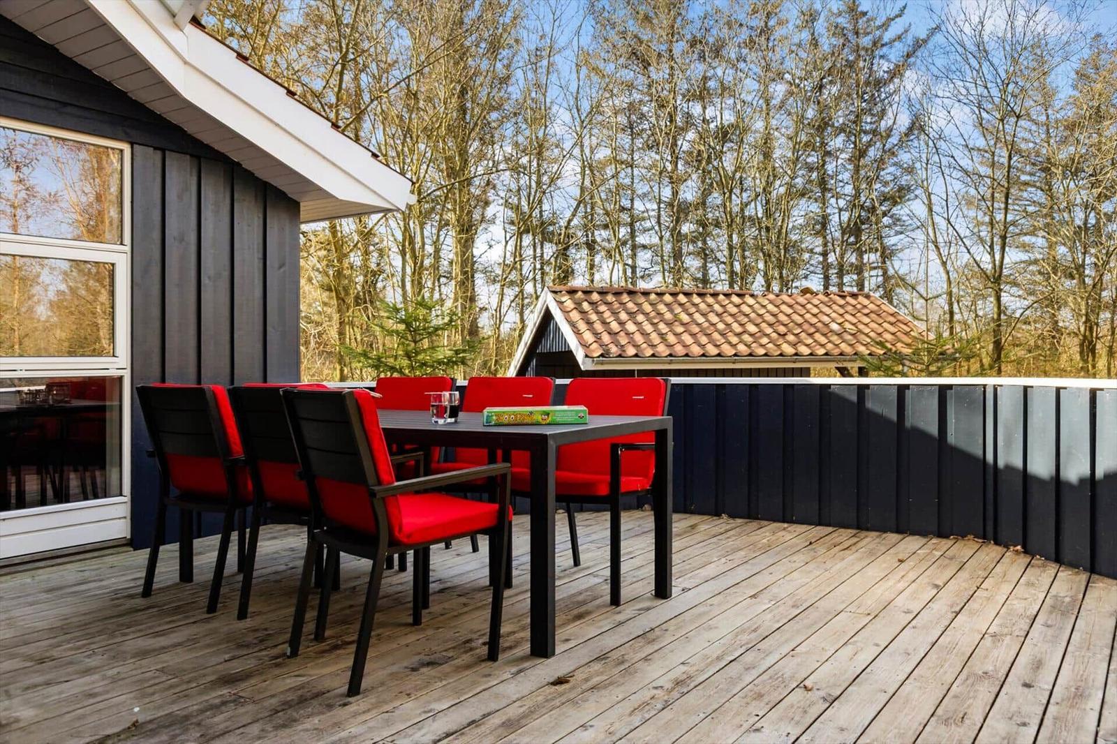 Terrasse mit Tisch und roten Stühlen, umgeben von Wald und Haus.
