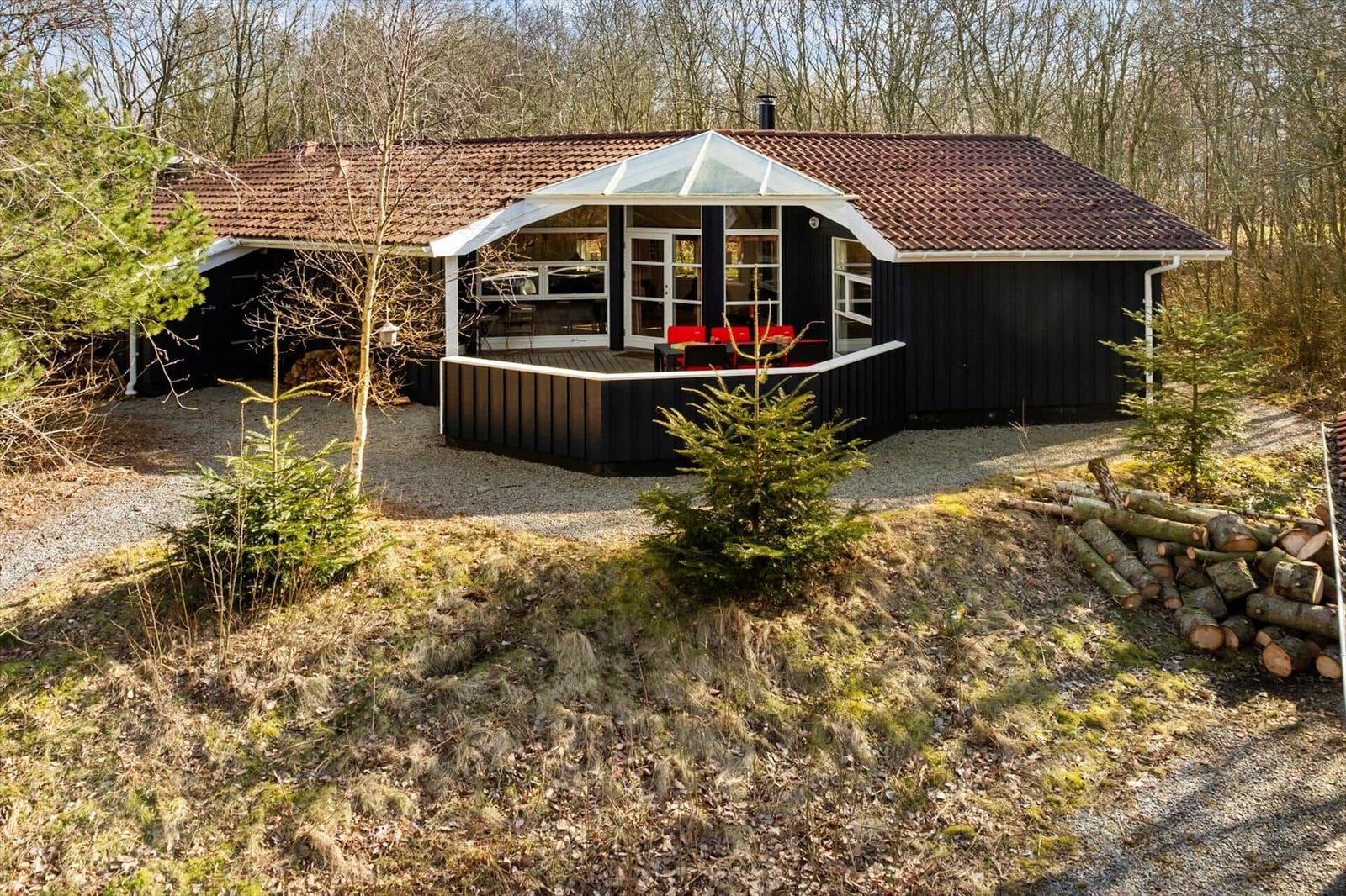 Schwarze Holzhütte mit Terrasse und rotem Dach, umgeben von Bäumen und einem Holzstapel.