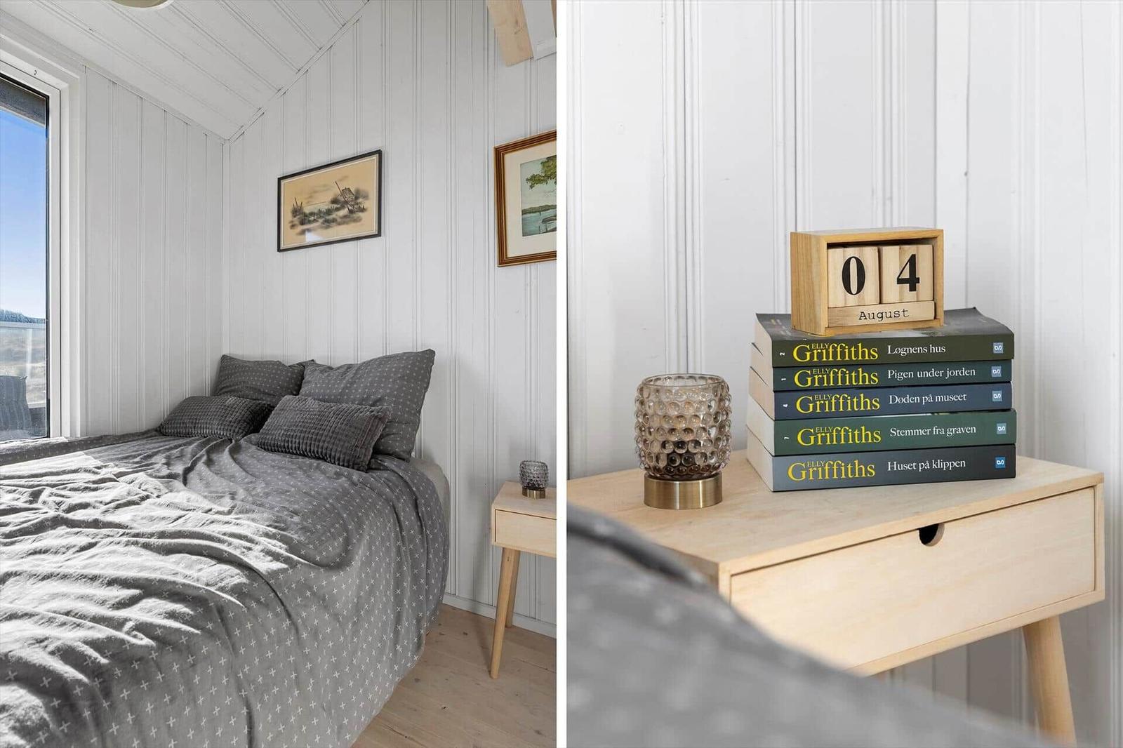 Schlafzimmer mit Bett, Nachttisch und Bücherstapel. Fenster zeigt Außenansicht.