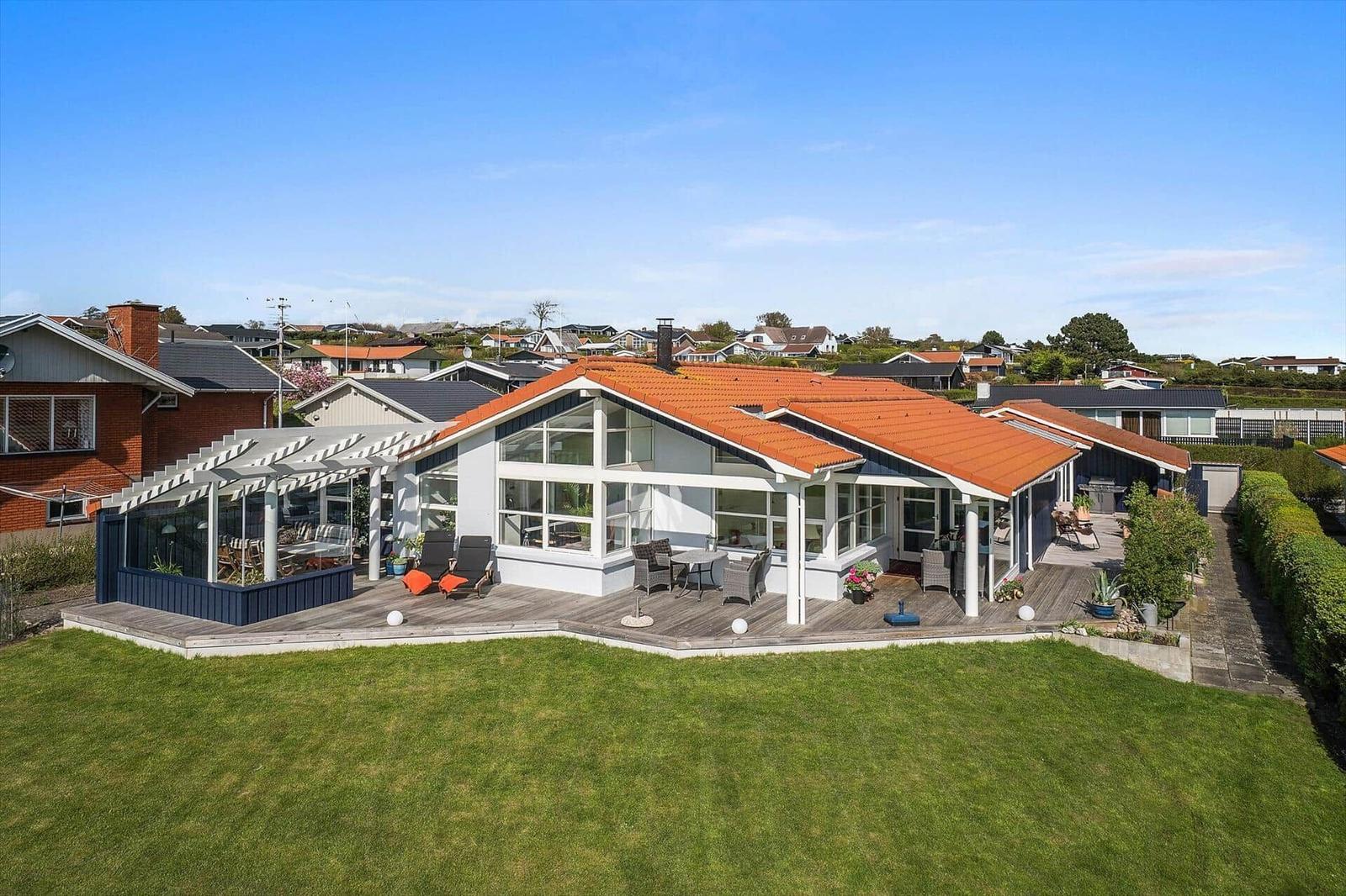 Modernes Haus mit großem Holzterrasse und rotem Dach. Grünfläche und Gartenbereich.