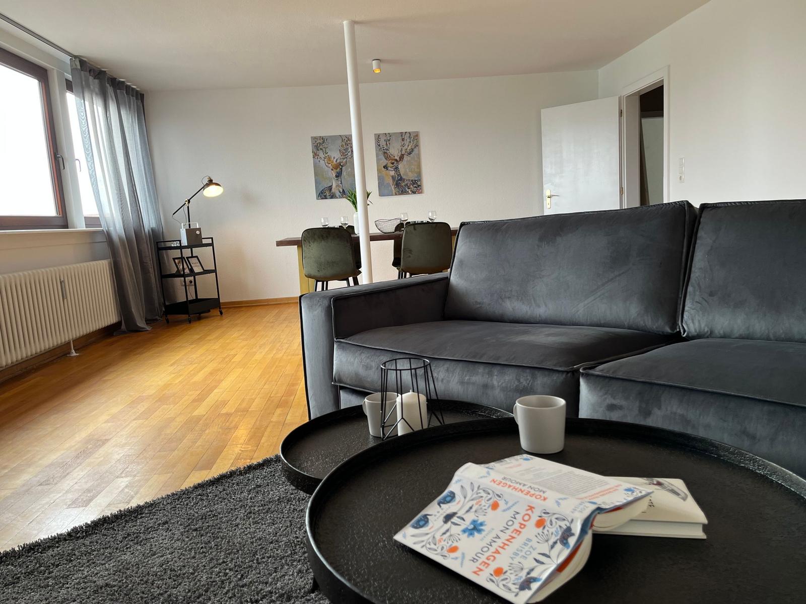 Wohnzimmer mit Sofa, Tisch und Fenster. Hintergrund zeigt Essbereich mit Bildern.