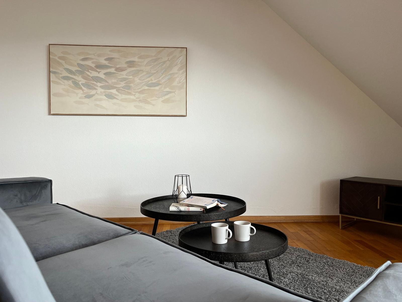 Ein Wohnbereich mit grauem Sofa, zwei runden Tischen und einem Wandbild.