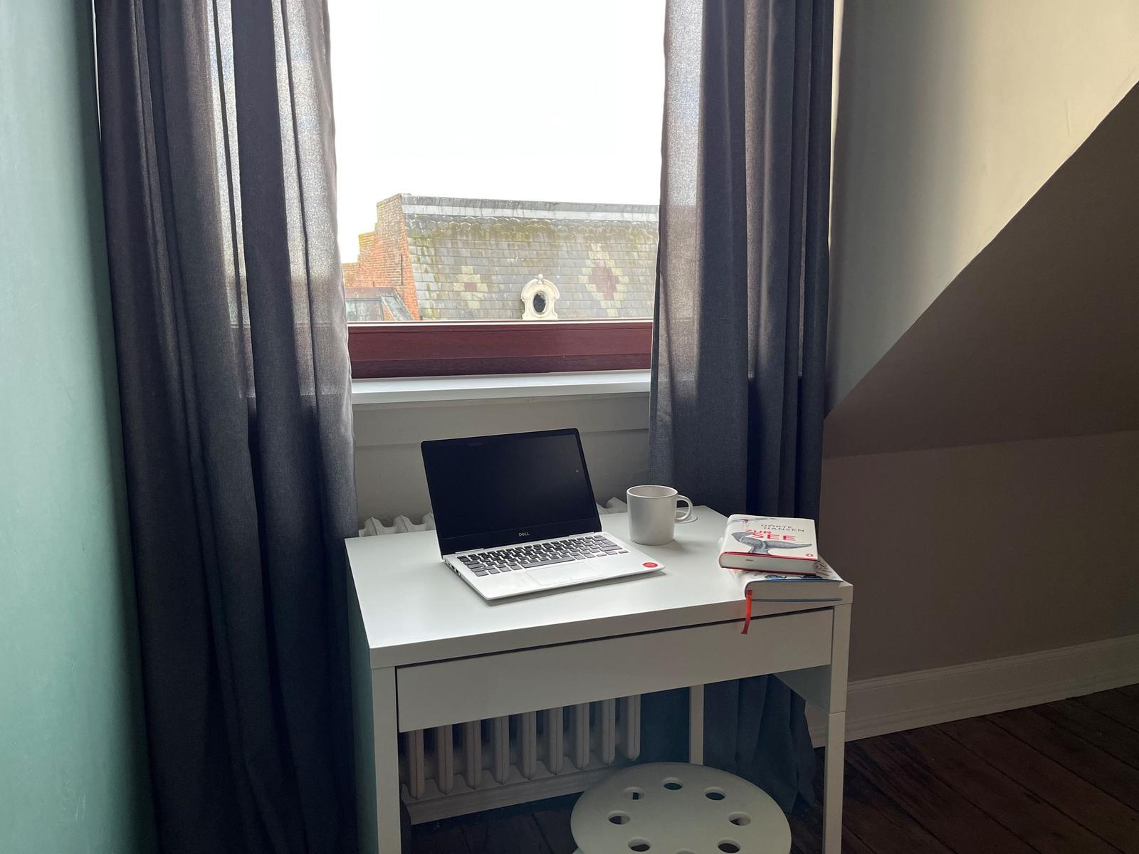 Schreibtisch mit Laptop, Tasse und Buch vor Fenster mit Blick auf Dach.