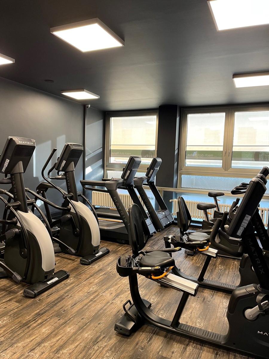 Fitnessraum mit Laufbändern, Stepper und Fahrrädern. Holzboden und Fenster.