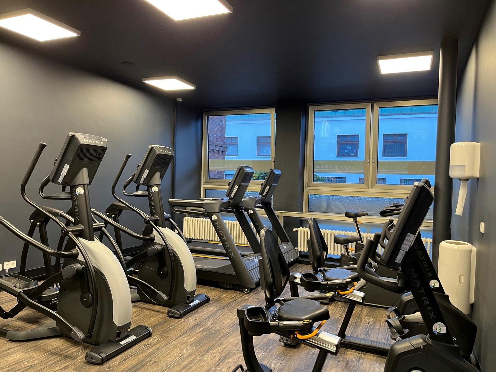 Fitnessraum mit Laufbändern und Crosstrainer, Fenster mit Blick auf Gebäude.