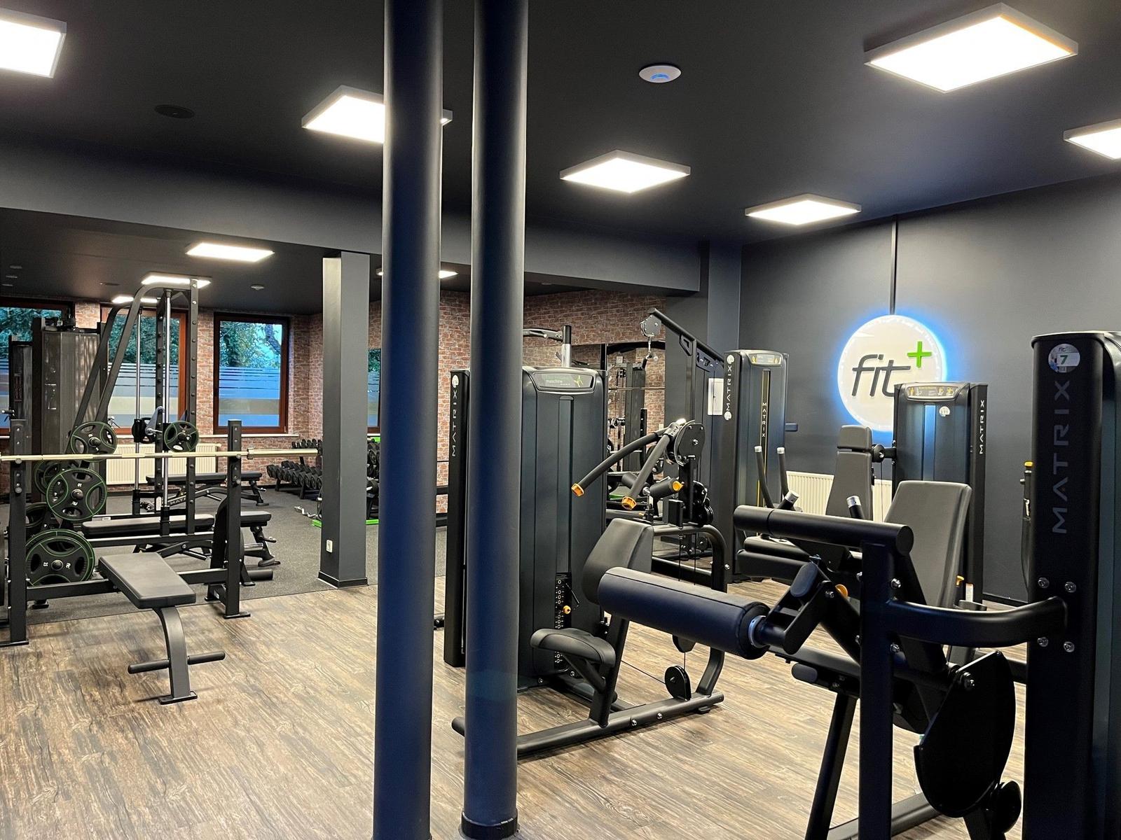 Fitnessraum mit Geräten und Holzboden. Wand mit Logo. Fenster im Hintergrund.
