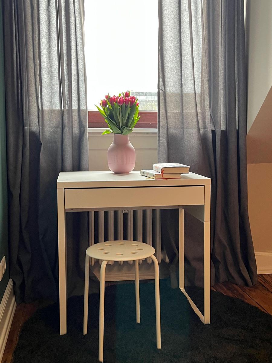 Schreibtisch mit Stuhl, Vase mit Tulpen und Büchern vor Fenster mit Vorhängen.